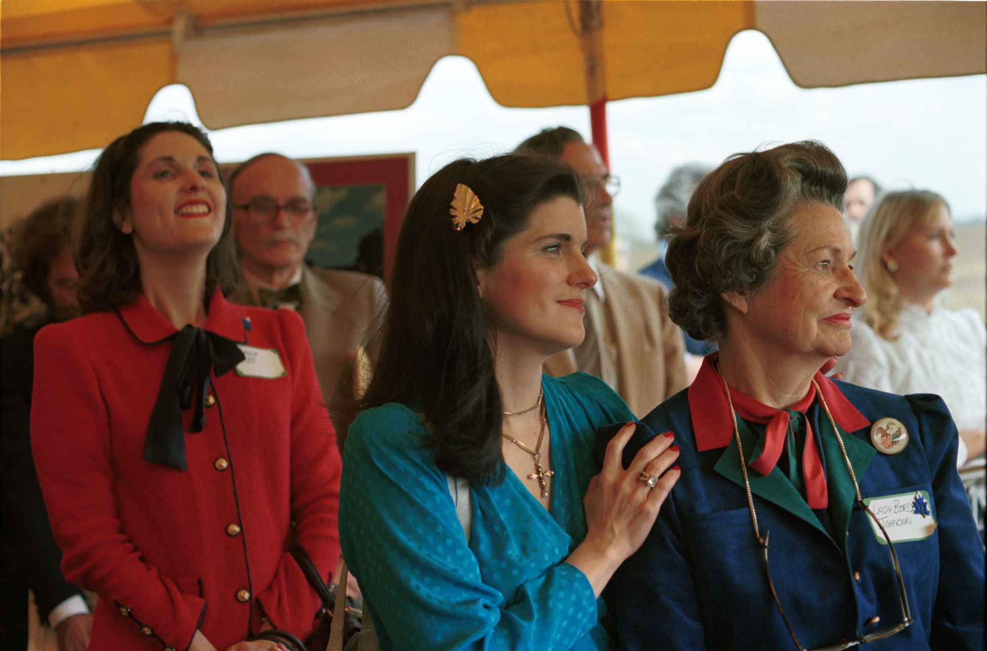 Lynda Johnson Robb in a red suit, Luci Baines Johnson in a teal blouse, and Lady Bird Johnson in a navy suit with red and green shirt under a tent, looking to their left.